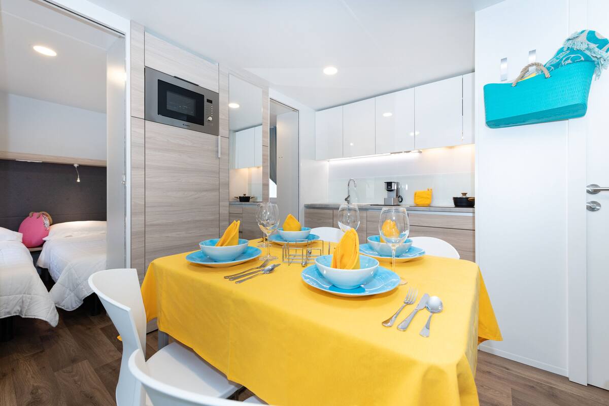 A modern hotel room with a dining area set for four, a kitchenette, and a bed with a pink suitcase.