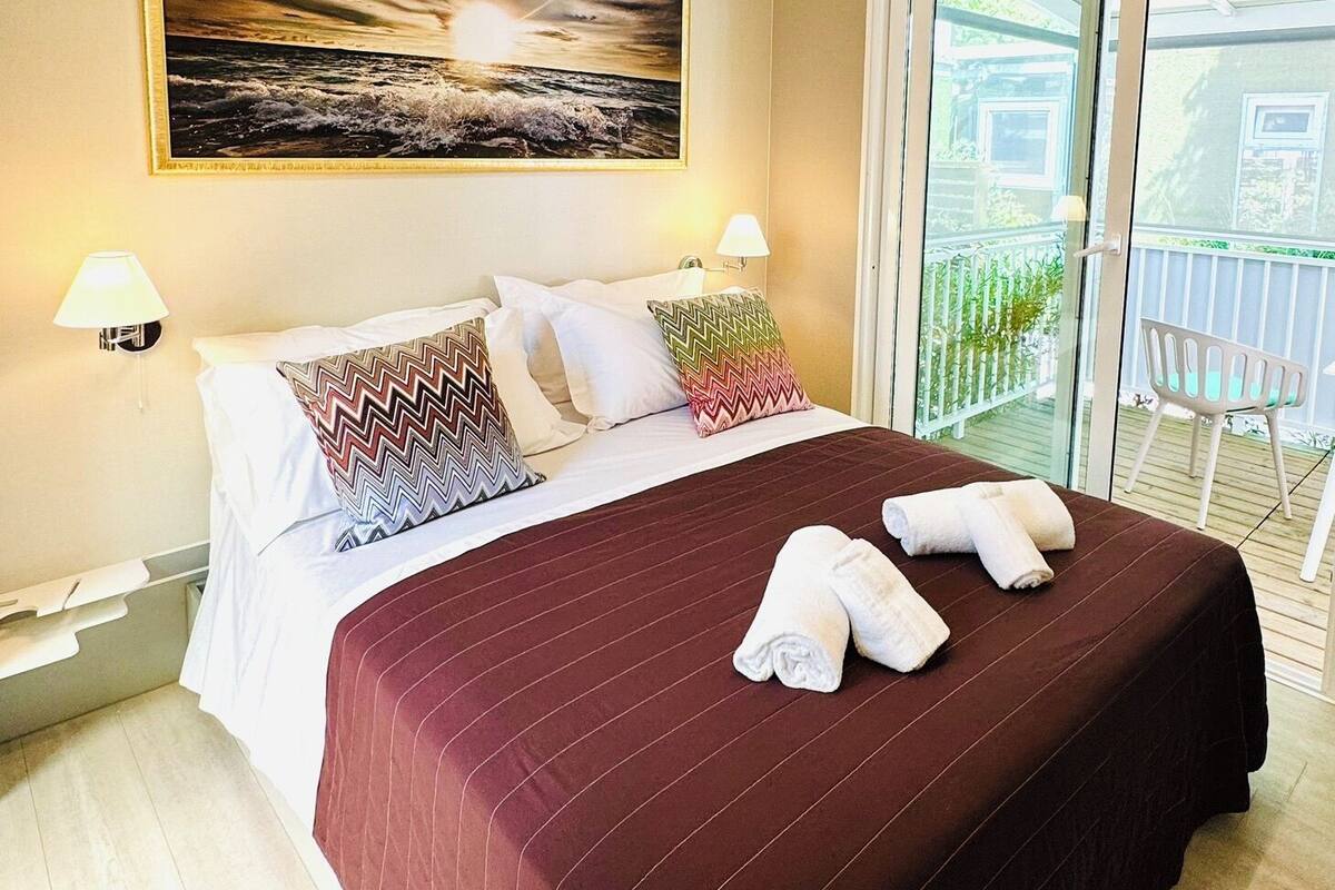 A hotel room with a large bed, a painting on the wall, and a balcony with a chair.