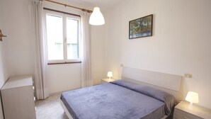 A bedroom with a bed, bedside tables, a window with curtains, a painting on the wall, and a wardrobe. - Appartamento Lungomare (Campo nell'Elba)