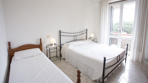 A bedroom with two single beds, a nightstand with a lamp, and a window with curtains. - Appartamento Lungomare (Campo nell'Elba)