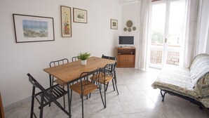 A room with a wooden dining table, four chairs, a small potted plant, a television on a wooden cabinet, a bed with patterned bedding, and a balcony with curtains. - Appartamento Lungomare (Campo nell'Elba)