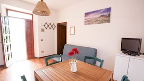 A dining area with a wooden table, green chairs, a white pitcher with flowers, a television on a cabinet, and a painting on the wall. - Appartamento Trilo La Padulella (Portoferraio)