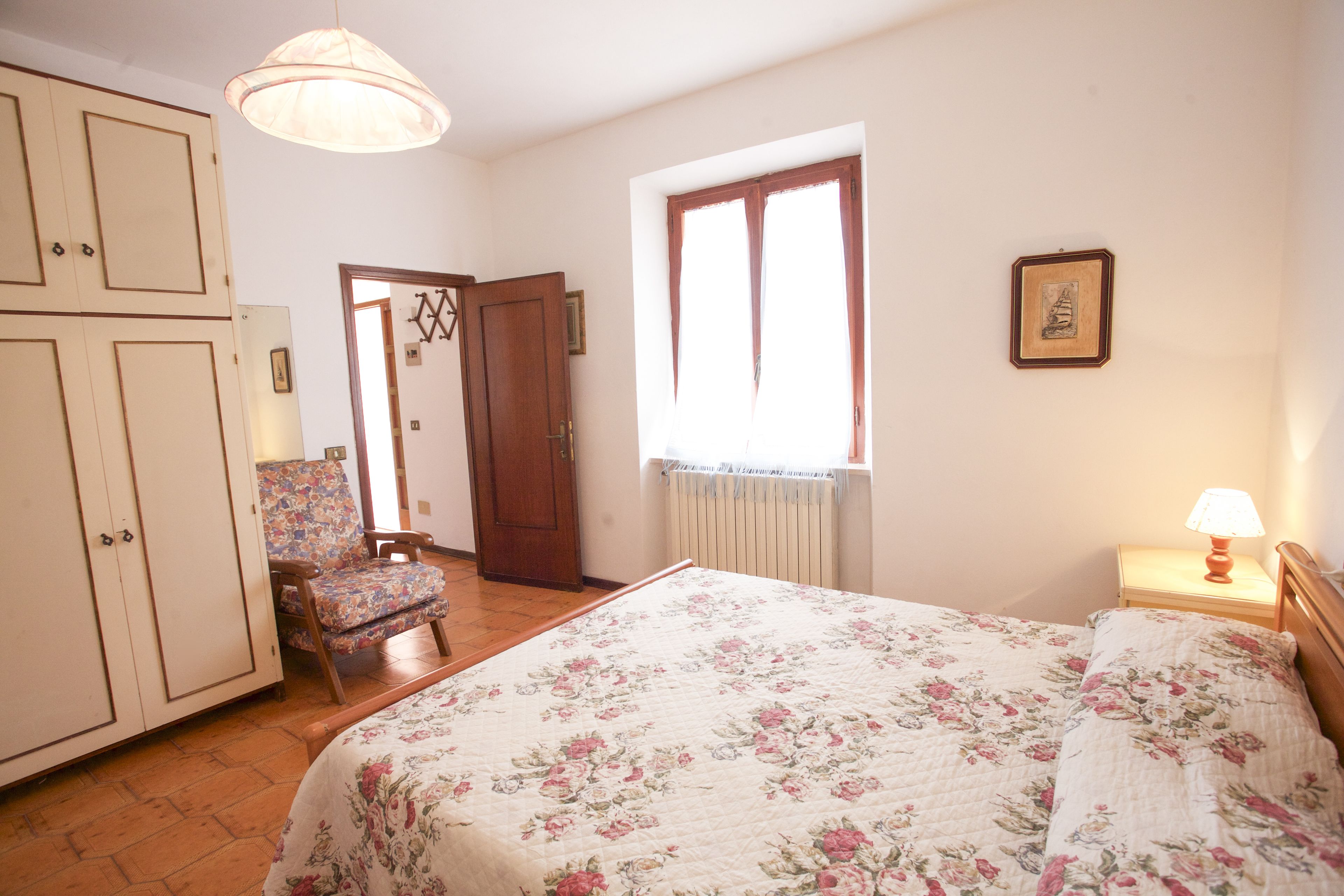 A bedroom with a floral bedspread, a wooden nightstand with a lamp, a chair, and a wardrobe.