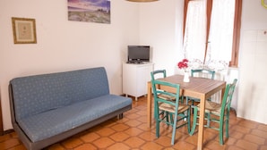A room with a blue sofa, a wooden dining table with four chairs, a small cabinet with a television, and a window with lace curtains. - Appartamento Trilo La Padulella (Portoferraio)