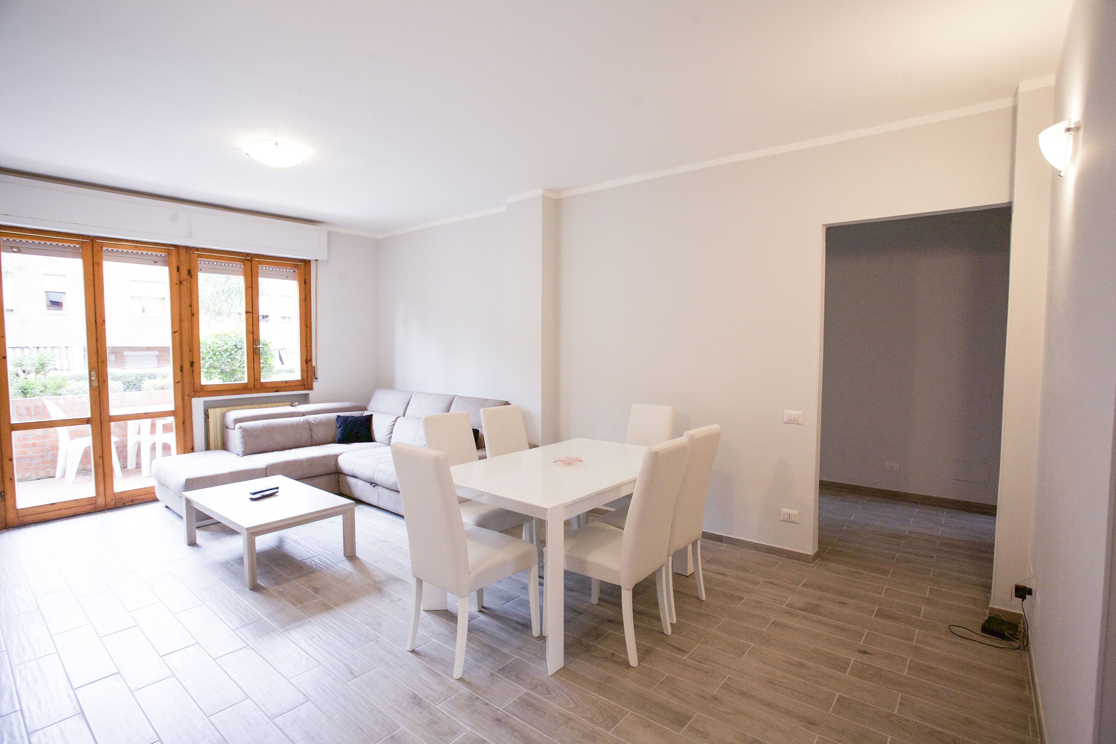 A modern living room with a white sofa, a dining table with chairs, and a large window with wooden frames.