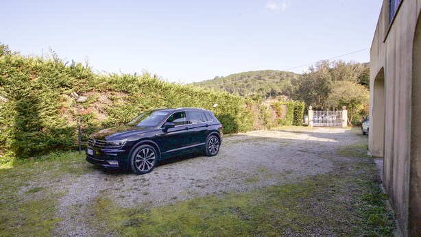A dark-colored car parked on a gravel driveway in a residential area with a house and a gate in the background. - Villino Mediterraneo (Portoferraio)