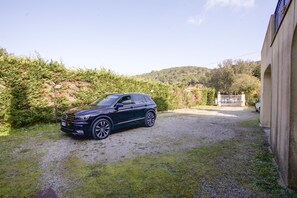 Un coche oscuro estacionado en un camino de grava en una zona residencial, con una casa y una puerta a lo lejos.