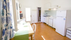 A compact kitchen with white appliances, a dining area with a green table, and a view into the adjacent room. - Appartamento il Mandarino (Portoferraio)