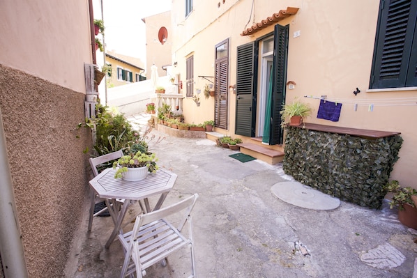 A narrow outdoor patio with a table and chairs, potted plants, and a stone wall.