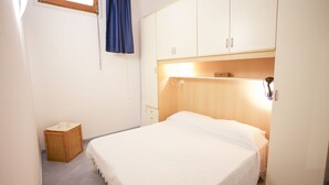A bedroom with a bed, a chest of drawers, a small wicker basket, and a window with a curtain.