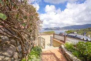Uma trilha costeira com um muro de pedra, um portão de metal e vista para o mar e montanhas.