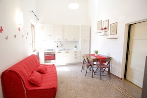 Un ambiente compatto con un divano rosso, un tavolo da pranzo con sedie, una zona cucina e una porta.