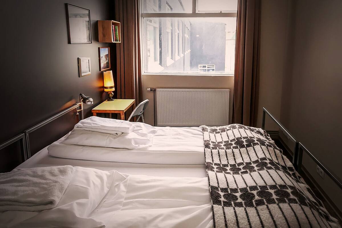 A hotel room with a bed, a desk, a chair, a lamp, a radiator, and a window with curtains.