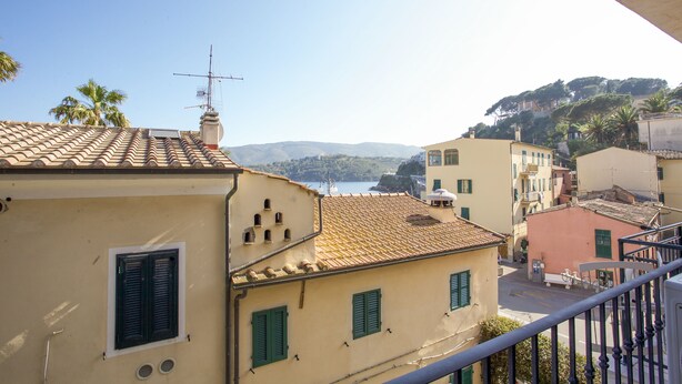 A coastal town with terracotta-roofed buildings, a clear sky, and a view of the sea and hills in the distance.