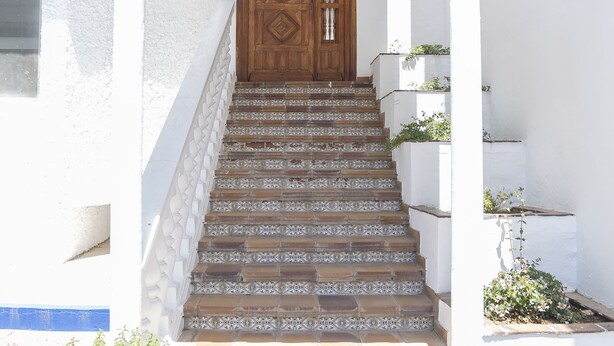 Una escalera conduce a una puerta de madera con un techo de azulejos decorativos, flanqueada por paredes blancas y macetas azules.
