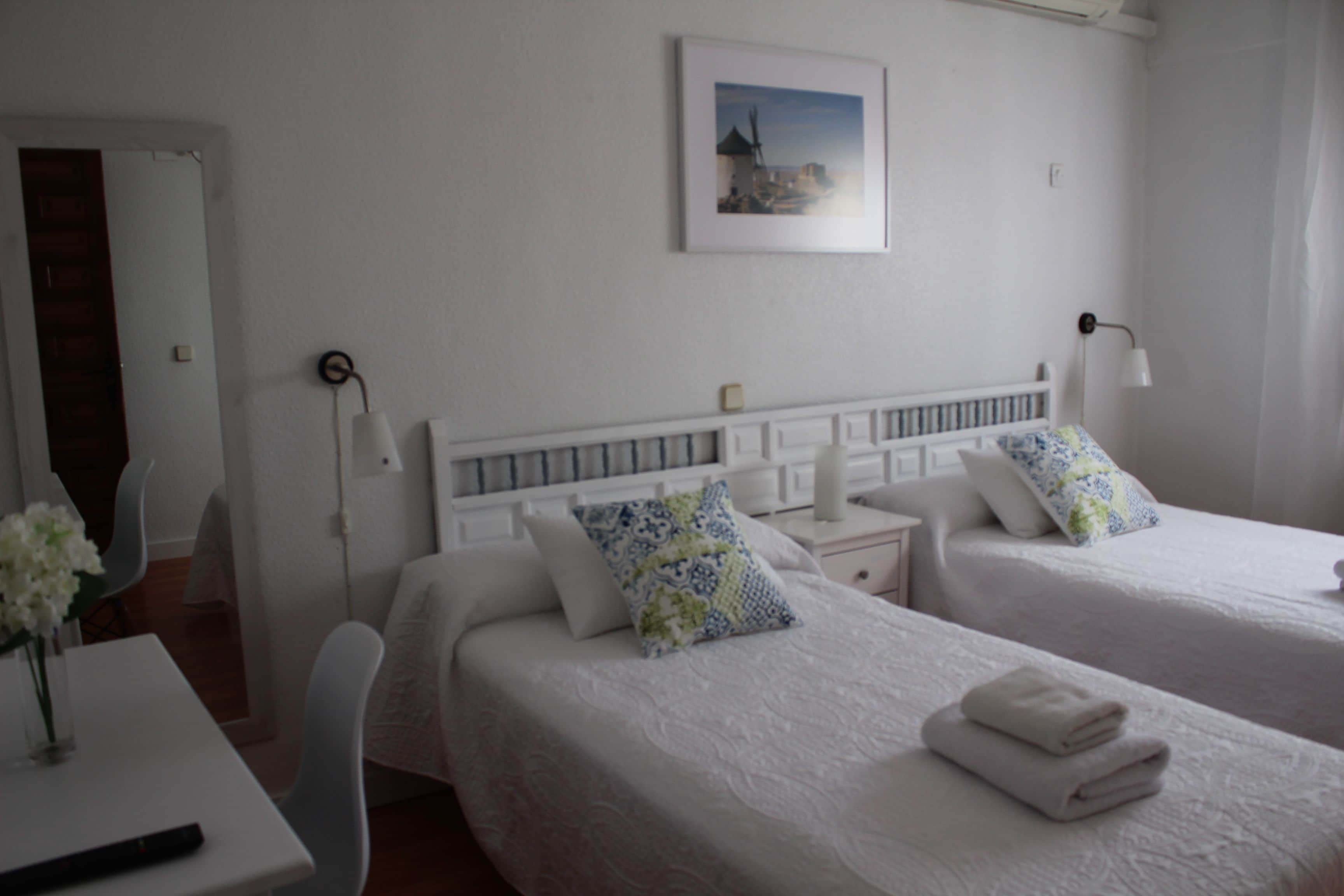 A hotel room with two beds, white bedding, a bedside table with a lamp, a chair, and a painting on the wall.