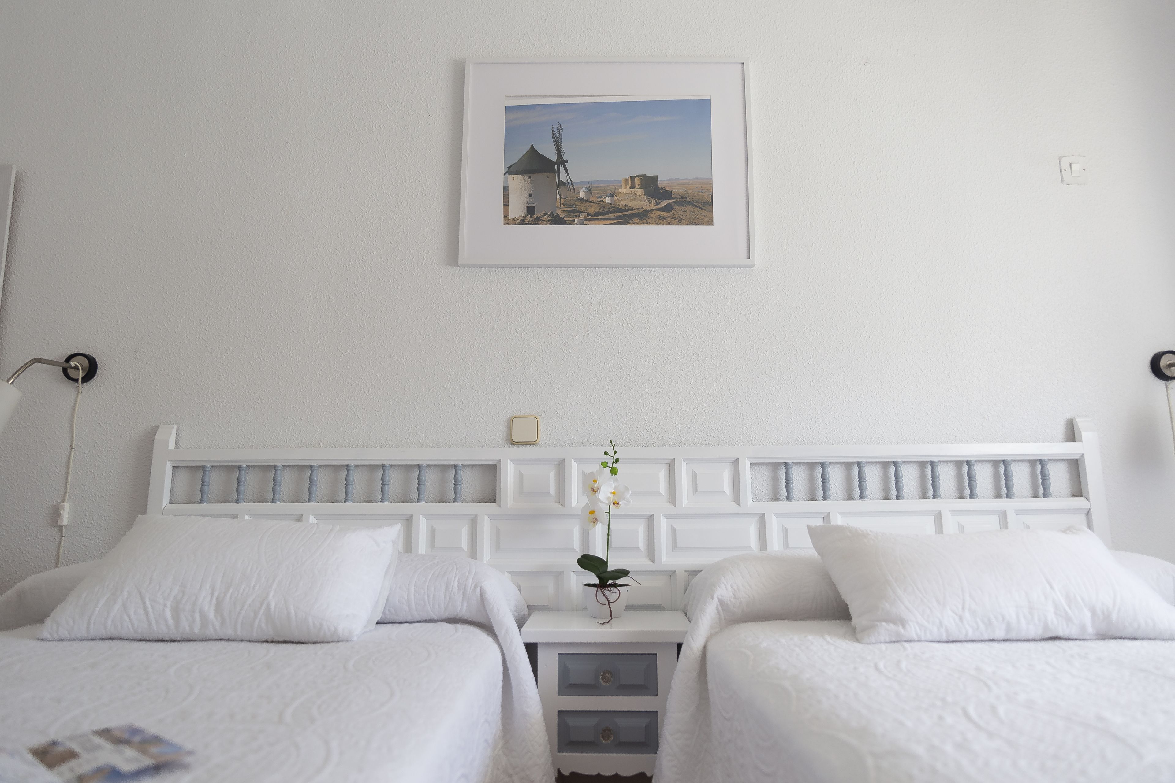 A hotel room with two beds, a white headboard, a framed picture on the wall, and a small table with a single orchid.