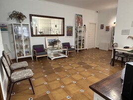 Uma sala de estar com piso xadrez, uma mesa de centro, duas poltronas, uma estante de livros e uma mesa de jantar com cadeiras.