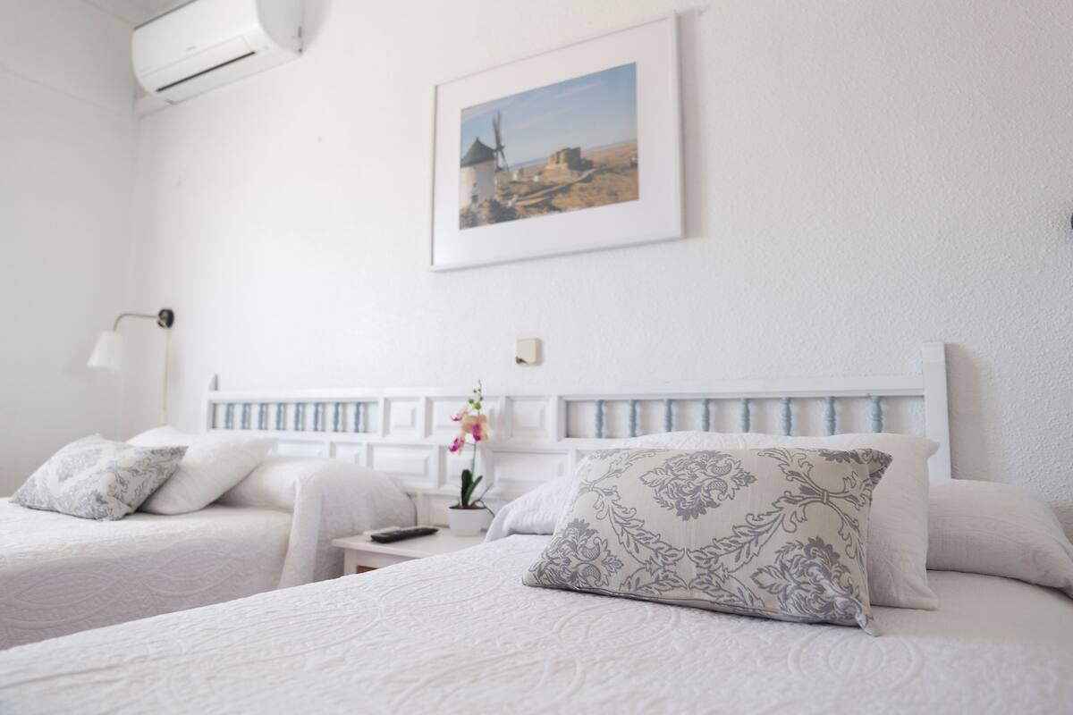 A hotel room with two beds, a white headboard, a framed picture on the wall, a bedside table with a lamp, and a potted plant.