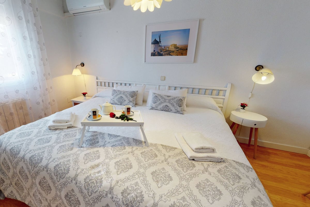 A neatly arranged bedroom with a bed, bedside tables, a bedside lamp, a picture on the wall, and a window with curtains.