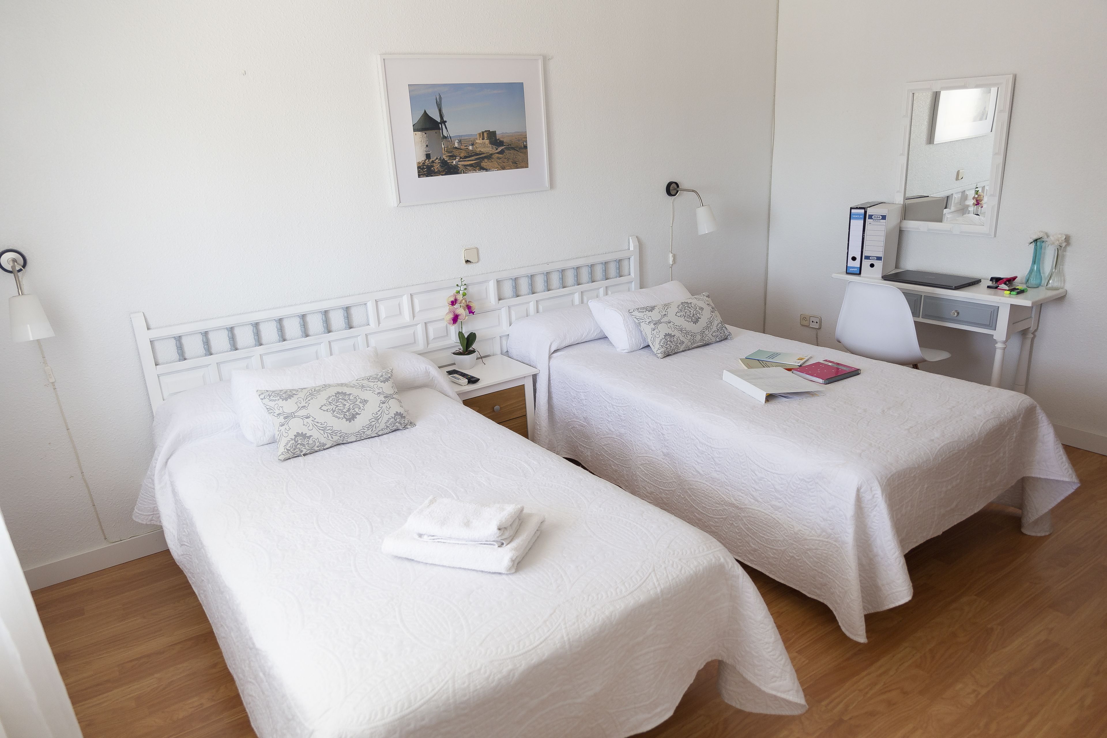 A hotel room with two single beds, a desk with a chair, a desk lamp, a mirror, and a framed picture on the wall.