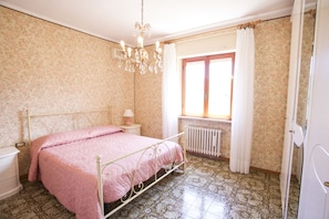 A bedroom with a bed, a nightstand, a dresser, a radiator, a window with curtains, and a chandelier. - Appartamento Il Portico (Portoferraio)