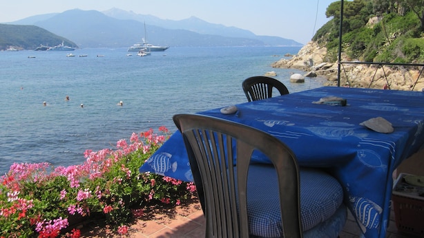 A table with a blue tablecloth and chairs, overlooking a body of water with boats and a mountainous backdrop. - Appartamento Il Sottomarino (Portoferraio)