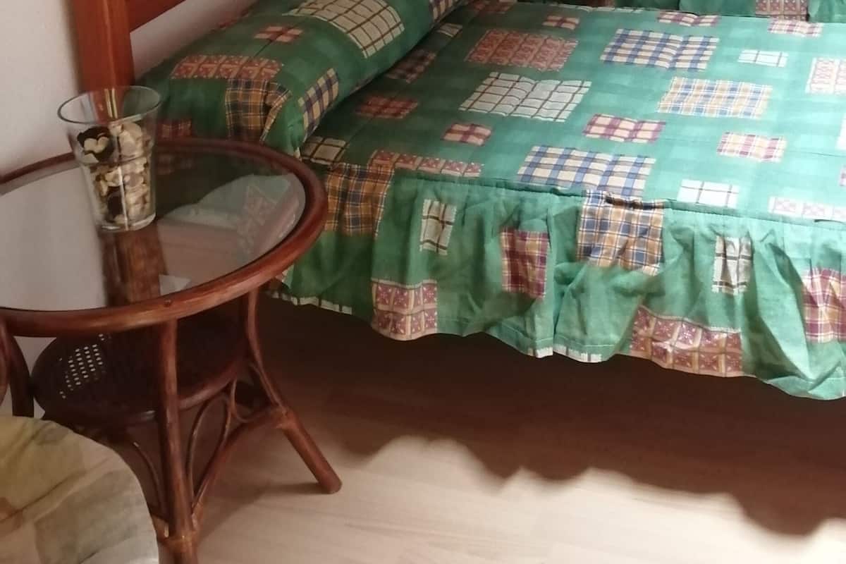 A small, cozy room with a wooden ceiling and a stone wall. It includes a bed with a green and white checkered bedspread, a wooden headboard, and a small round table with a glass top.