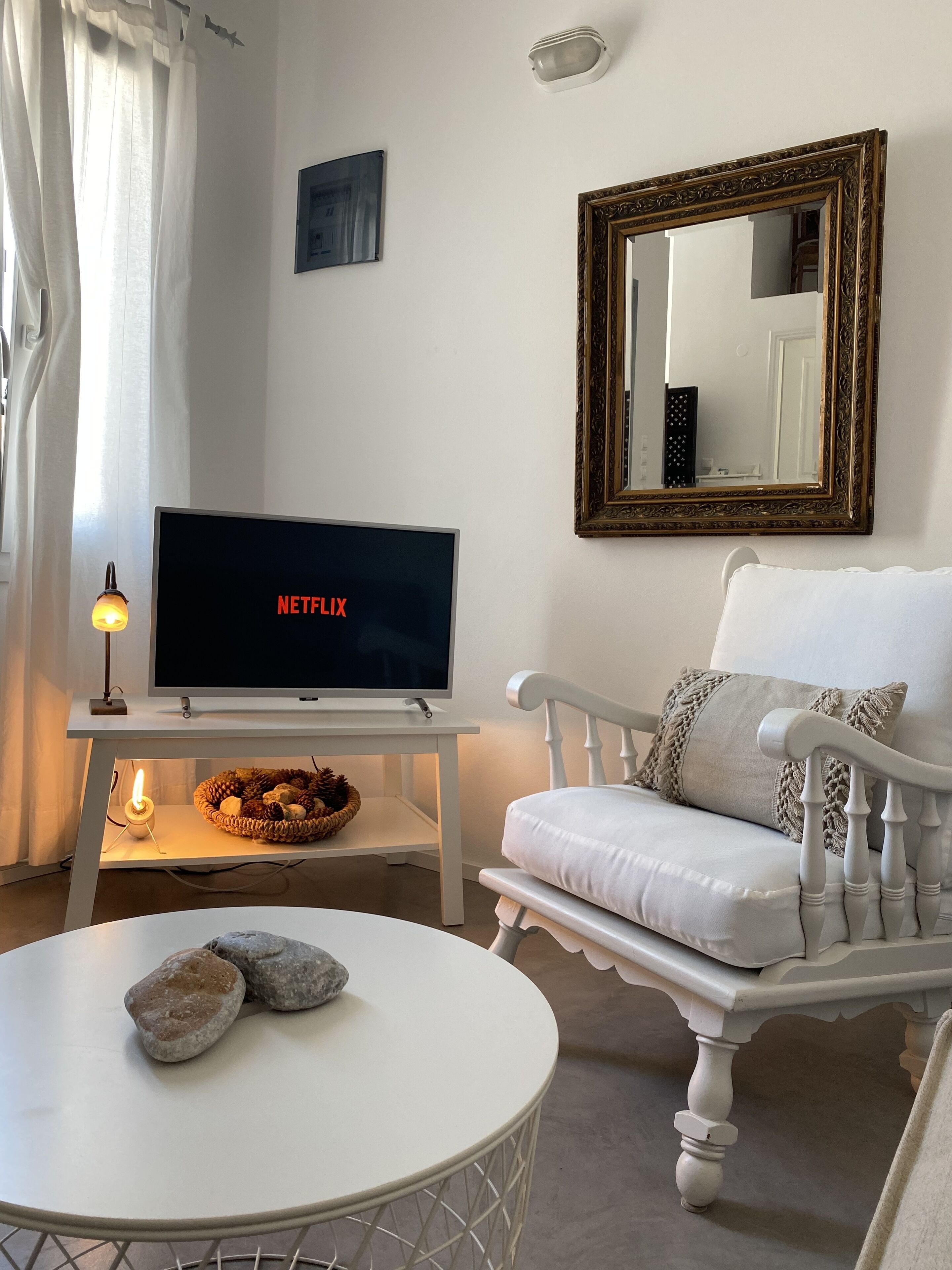Una sala de estar acogedora con un sofá blanco, una mesa redonda de centro con rocas, un televisor mostrando 'NETFLIX', un espejo decorativo y un aplique de pared.