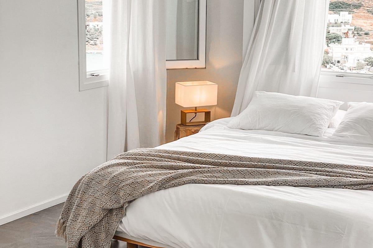 A bedroom with a bed, a bedside lamp, a window with curtains, and a view of a hilly landscape.