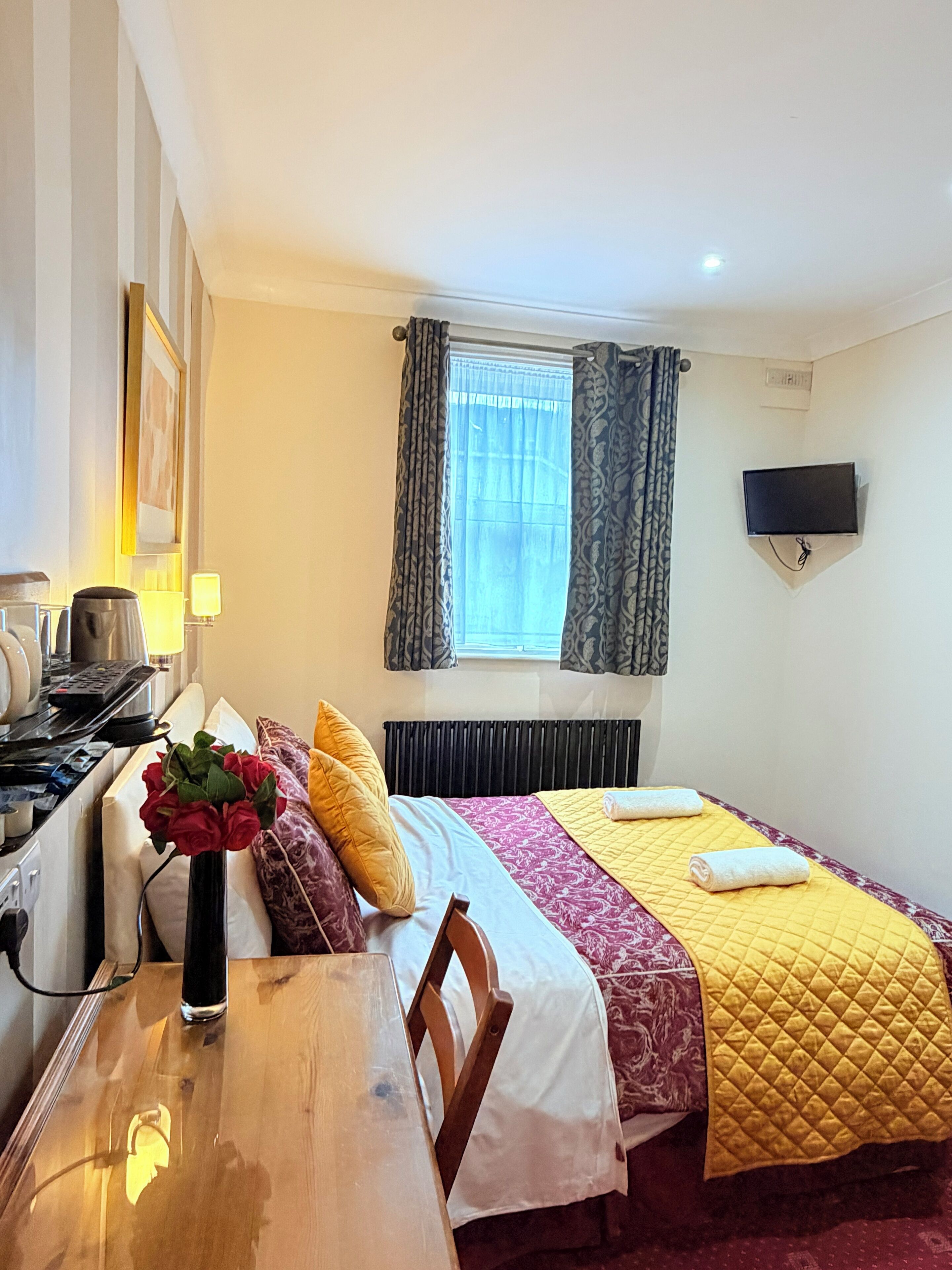 A hotel room with a bed, a TV mounted on the wall, a window with curtains, a radiator, a table with a vase of flowers, and a wooden chair.