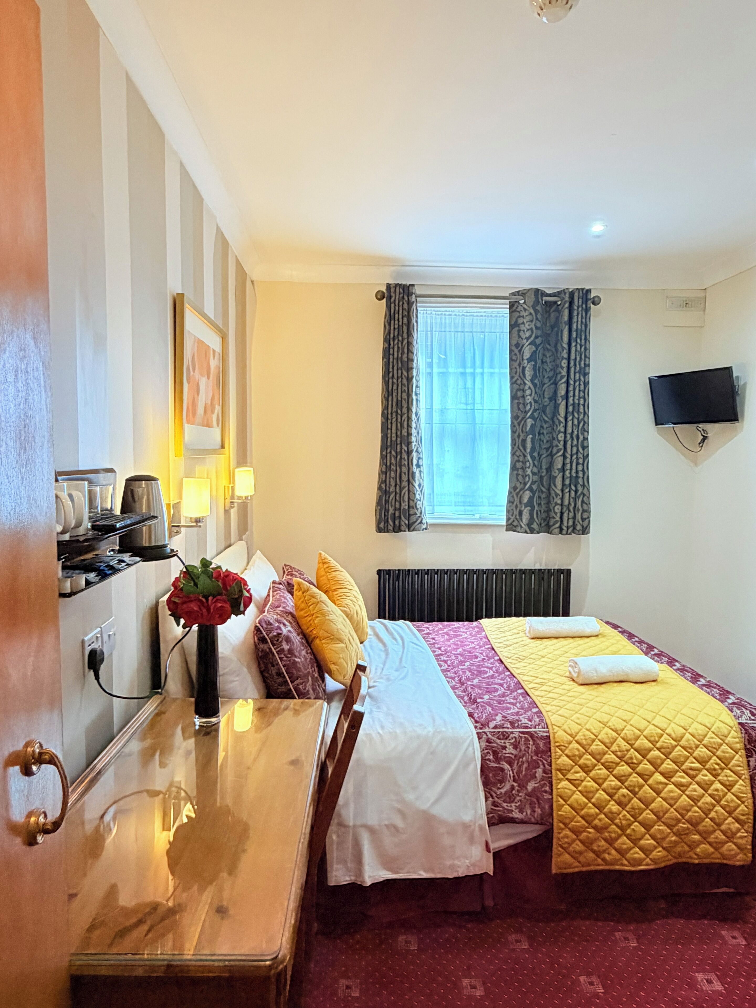Een hotelkamer met een bed, nachtkastje, lamp en een aan de muur bevestigde televisie.