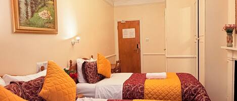 Habitación de hotel con tres camas, cada una con ropa de cama estampada y almohadas amarillas. Hay una araña y un cuadro en la pared, además de una puerta con un cartel.