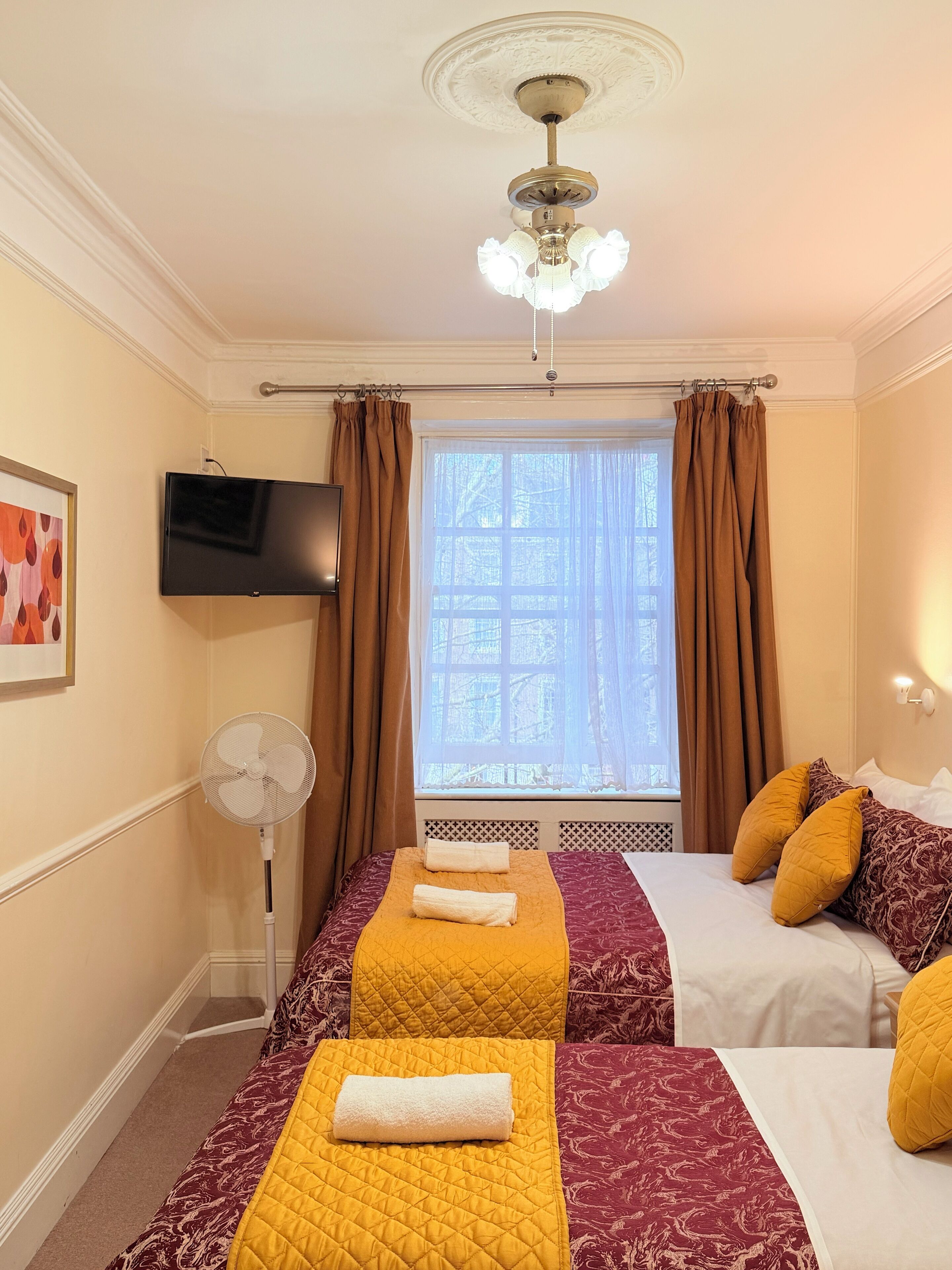 Habitación de hotel con dos camas, ventilador de techo, televisión y ventana con cortinas.