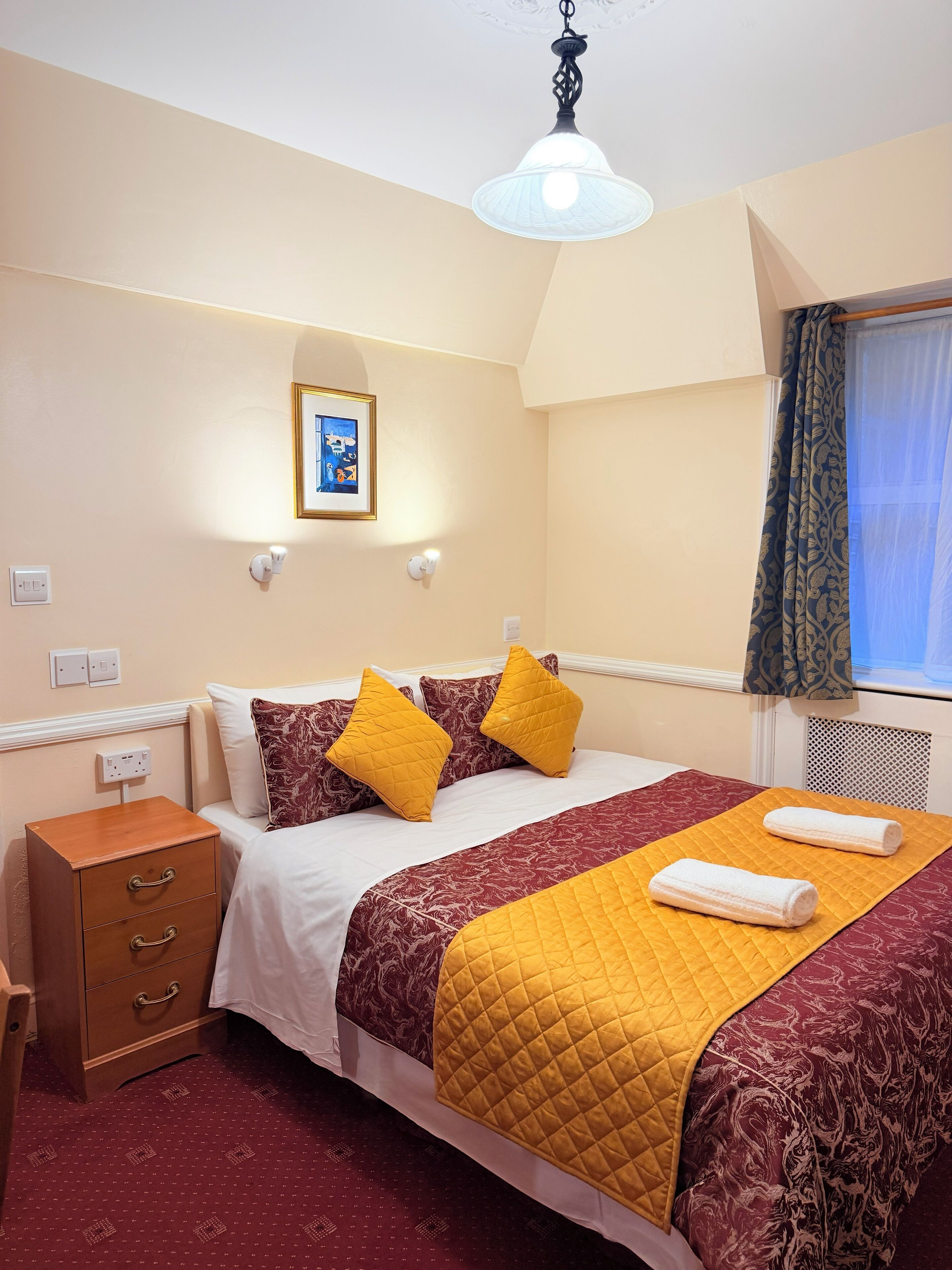A hotel room with a bed, a nightstand, a picture on the wall, and a window with curtains.