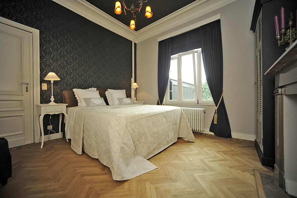 A bedroom with a large bed, two bedside tables, a radiator, a window with curtains, and a chandelier.