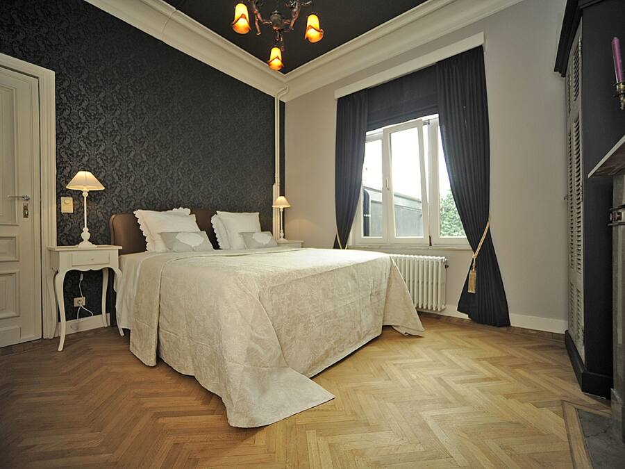 Ein Schlafzimmer mit einem großen Bett, zwei Nachttischen, einem Heizkörper, einem Fenster mit Vorhängen und einer Kristallleuchtre.