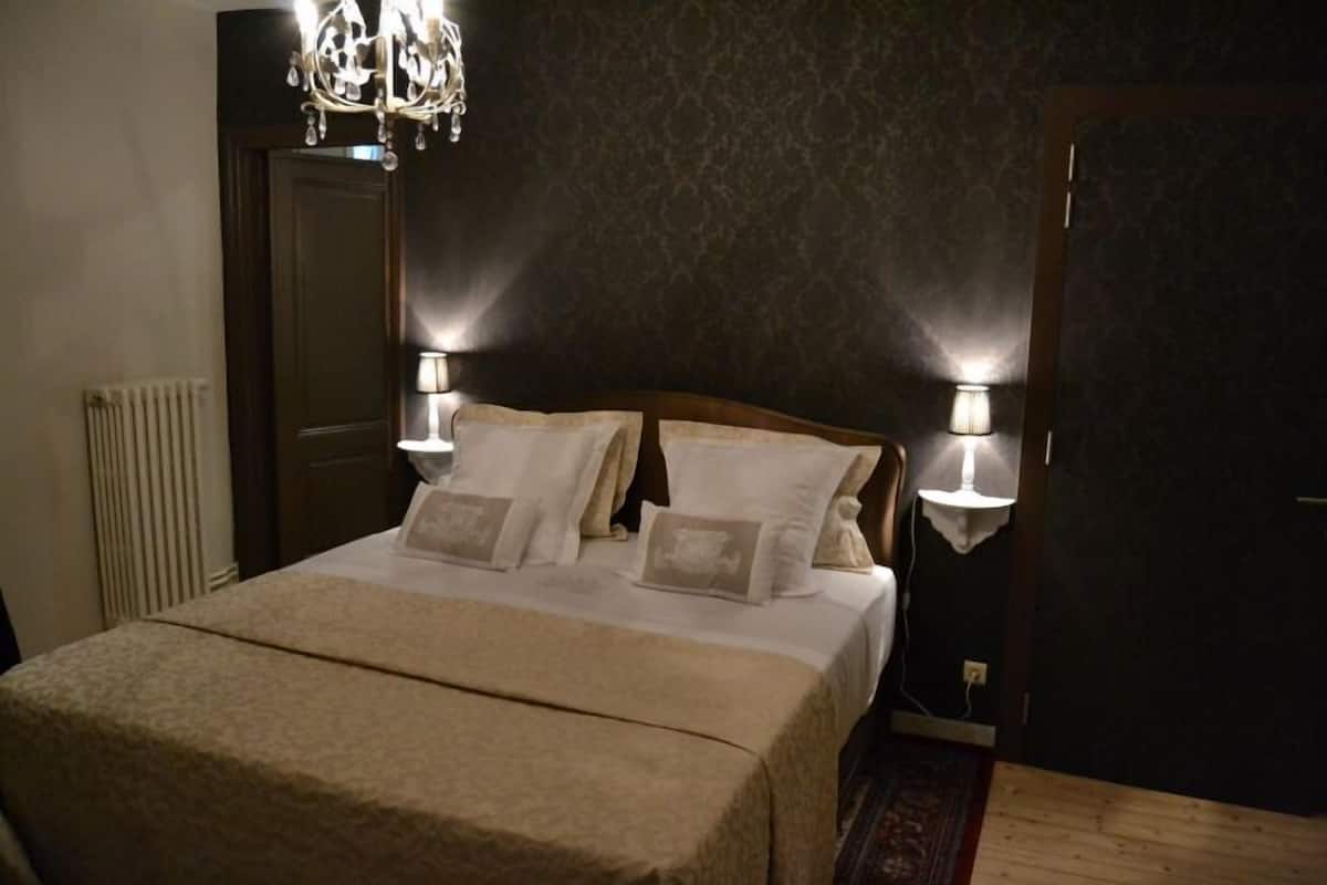 A neatly made bed with a beige bedspread and pillows, two bedside lamps, a chandelier, and a dark patterned wall.