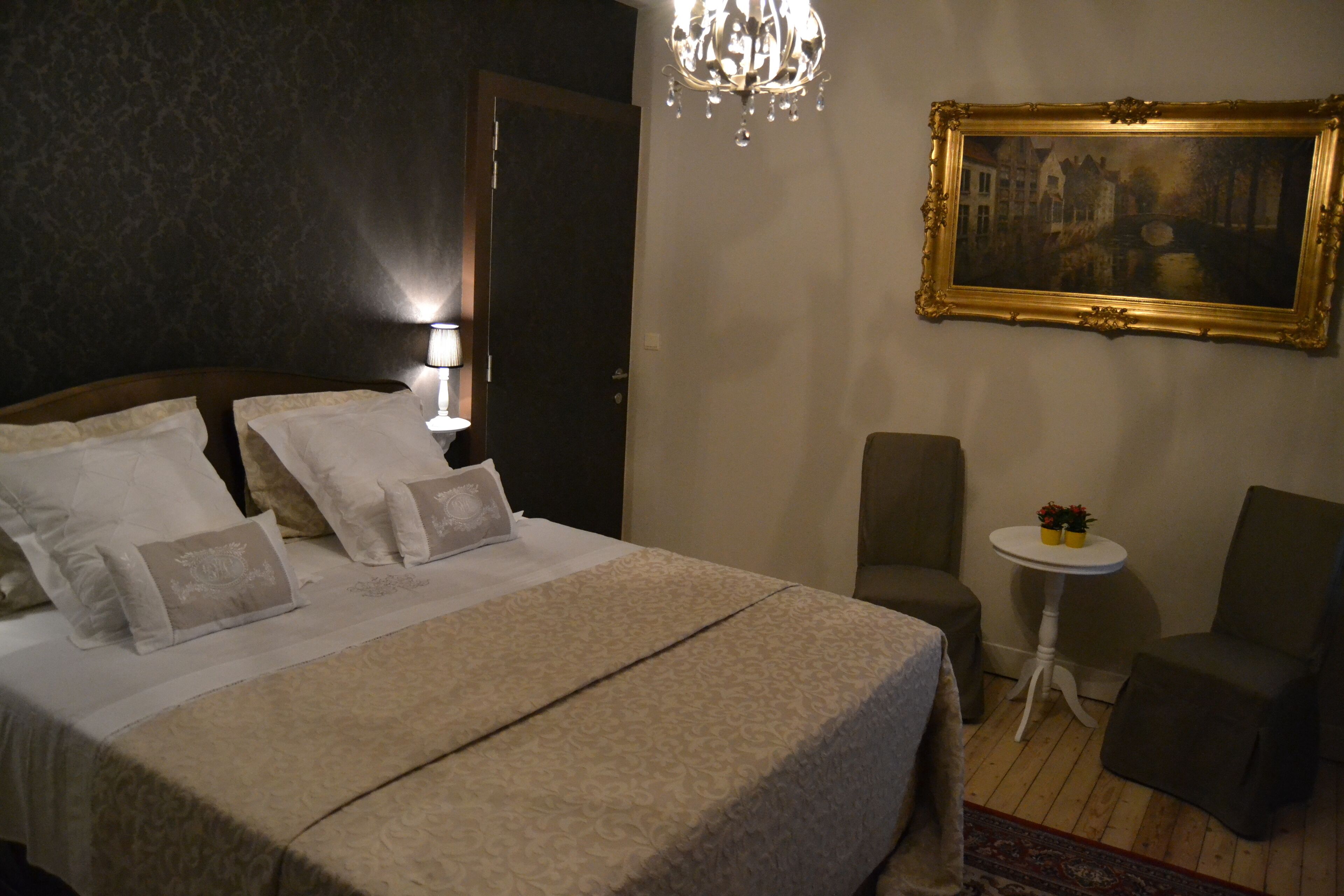 A hotel room with a large bed, a chandelier, a framed painting, a small table, and two chairs.