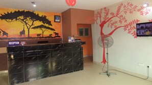 A reception area with a black marble counter, a reception sign, a wall mural of a savannah scene, a red heart decoration, and a wall-mounted television. - DC Bar & Suites (Lagos)
