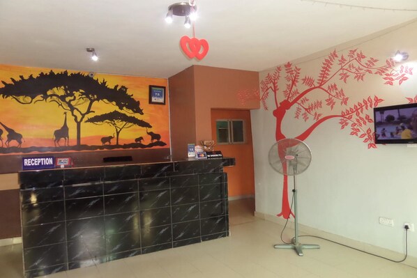 Un'area reception con un bancone in marmo nero, un cartello della reception, un affresco raffigurante una savana, una decorazione a forma di cuore rosso e un televisore fissato alla parete.