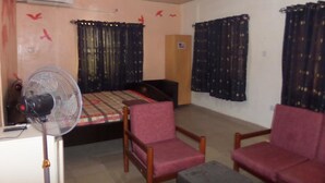 A room with a bed, two chairs, a standing fan, and a door. - DC Bar & Suites (Lagos)