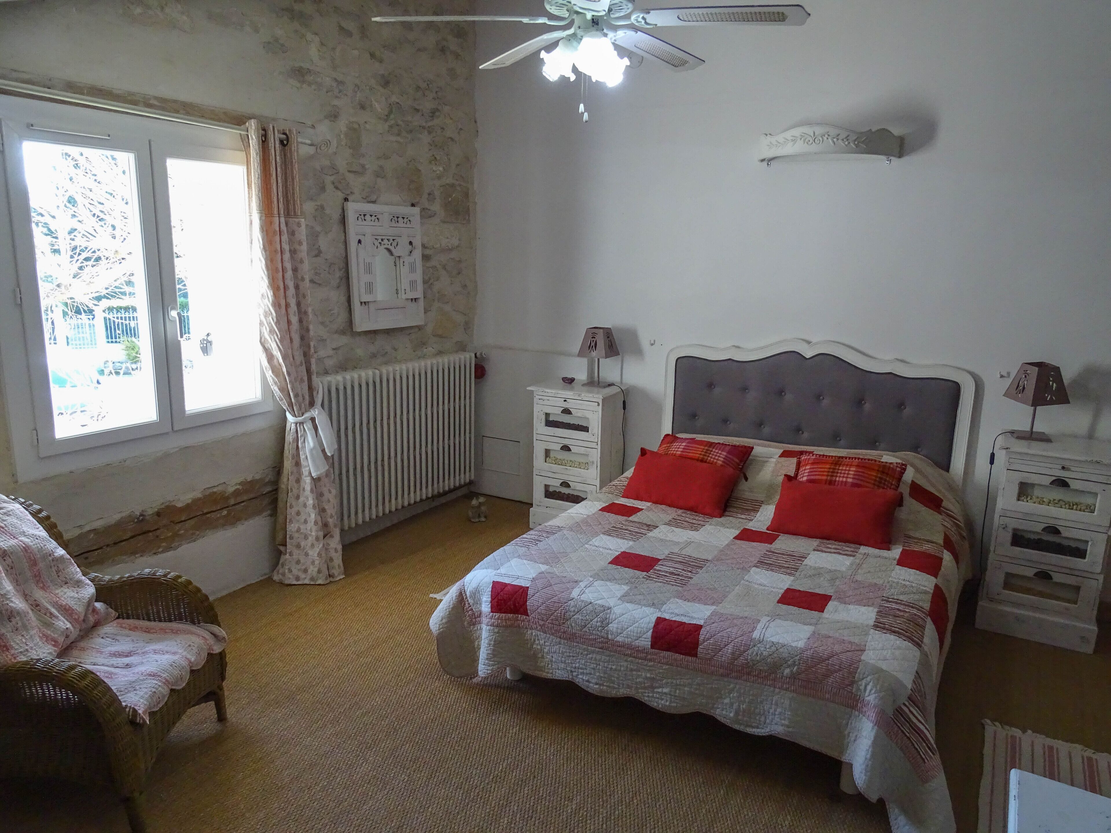 A bedroom with a bed, two bedside tables, a chair, a radiator, a window with curtains, and a ceiling fan.