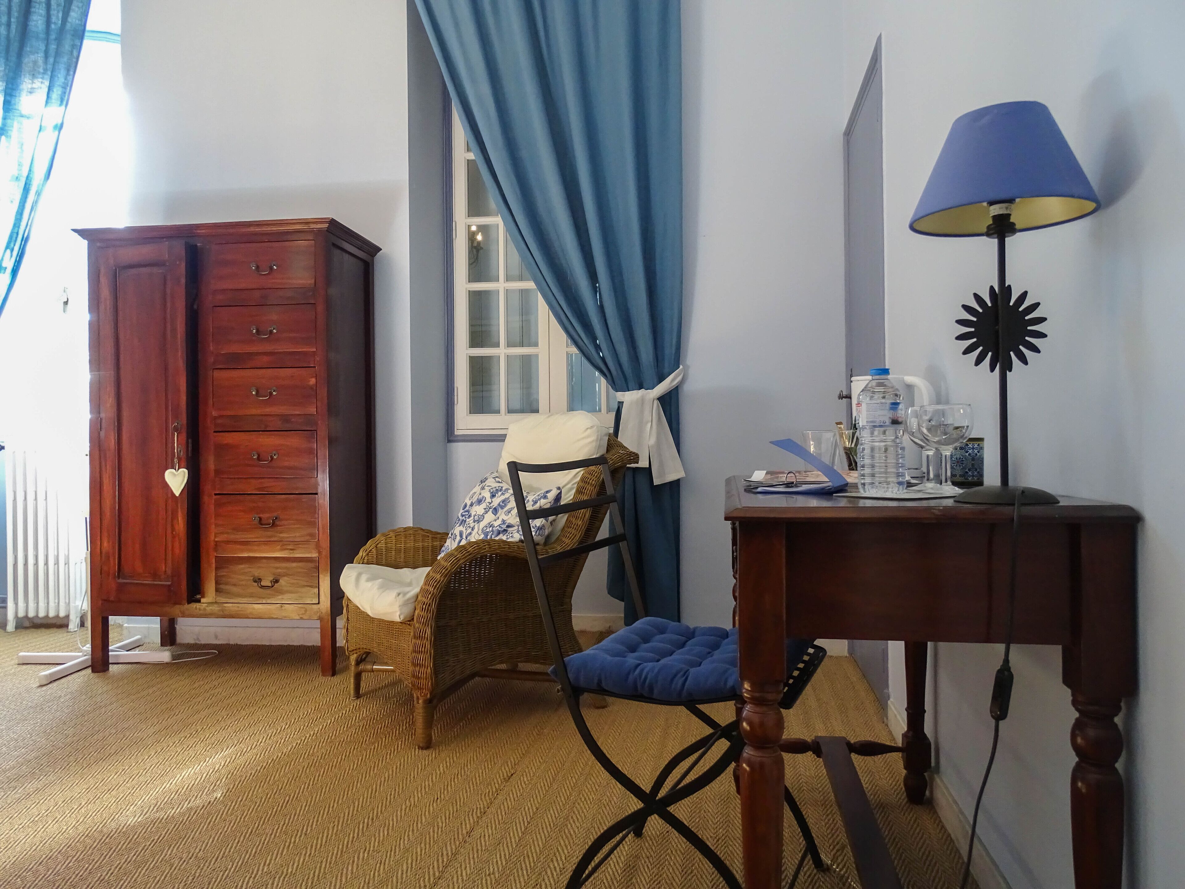 A room with a wooden cabinet, wicker chair, blue cushion, and a table with a lamp and water bottle.