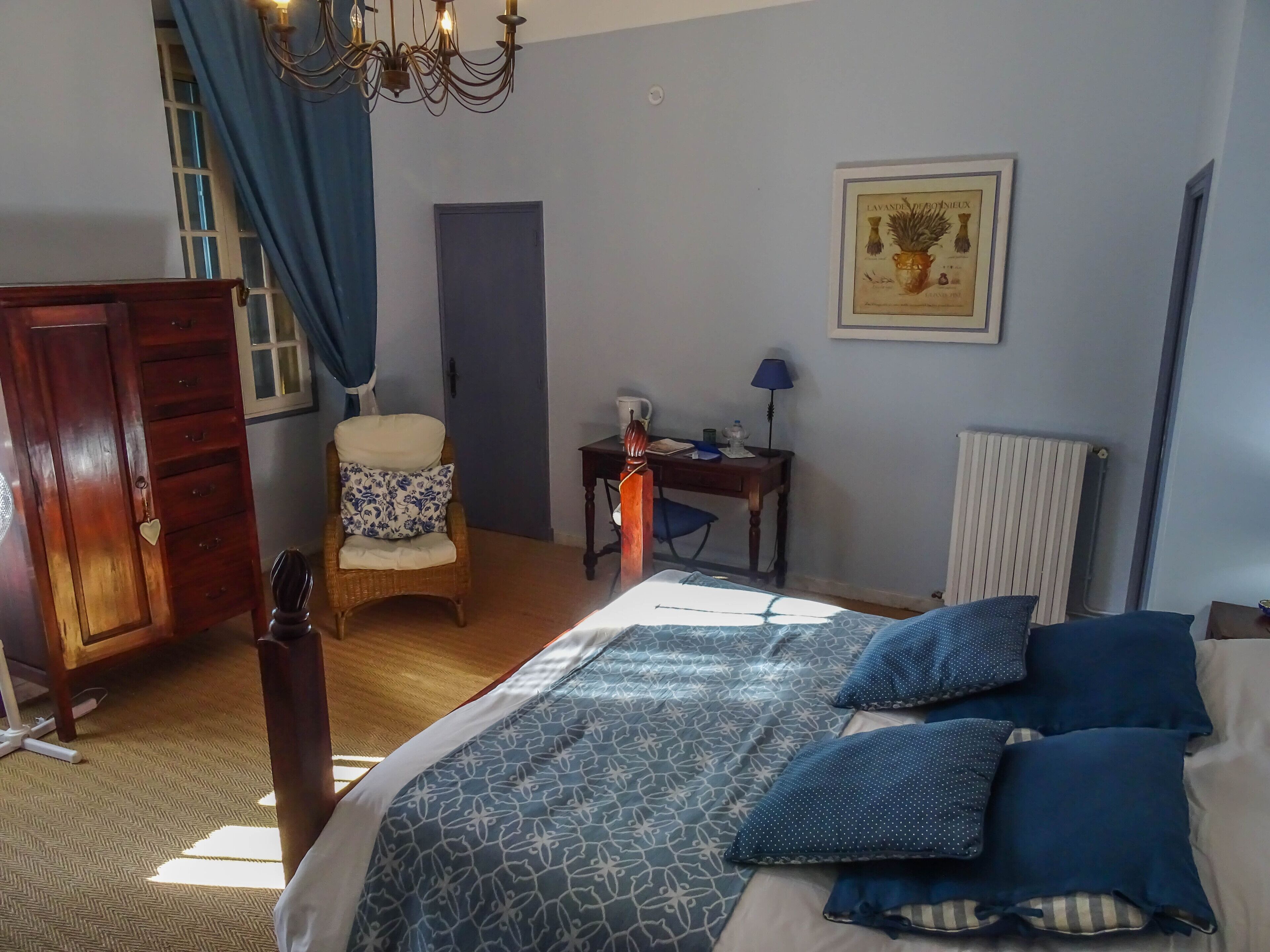 A bedroom with a bed, a wooden wardrobe, a chair, a desk, a radiator, and a framed picture on the wall.