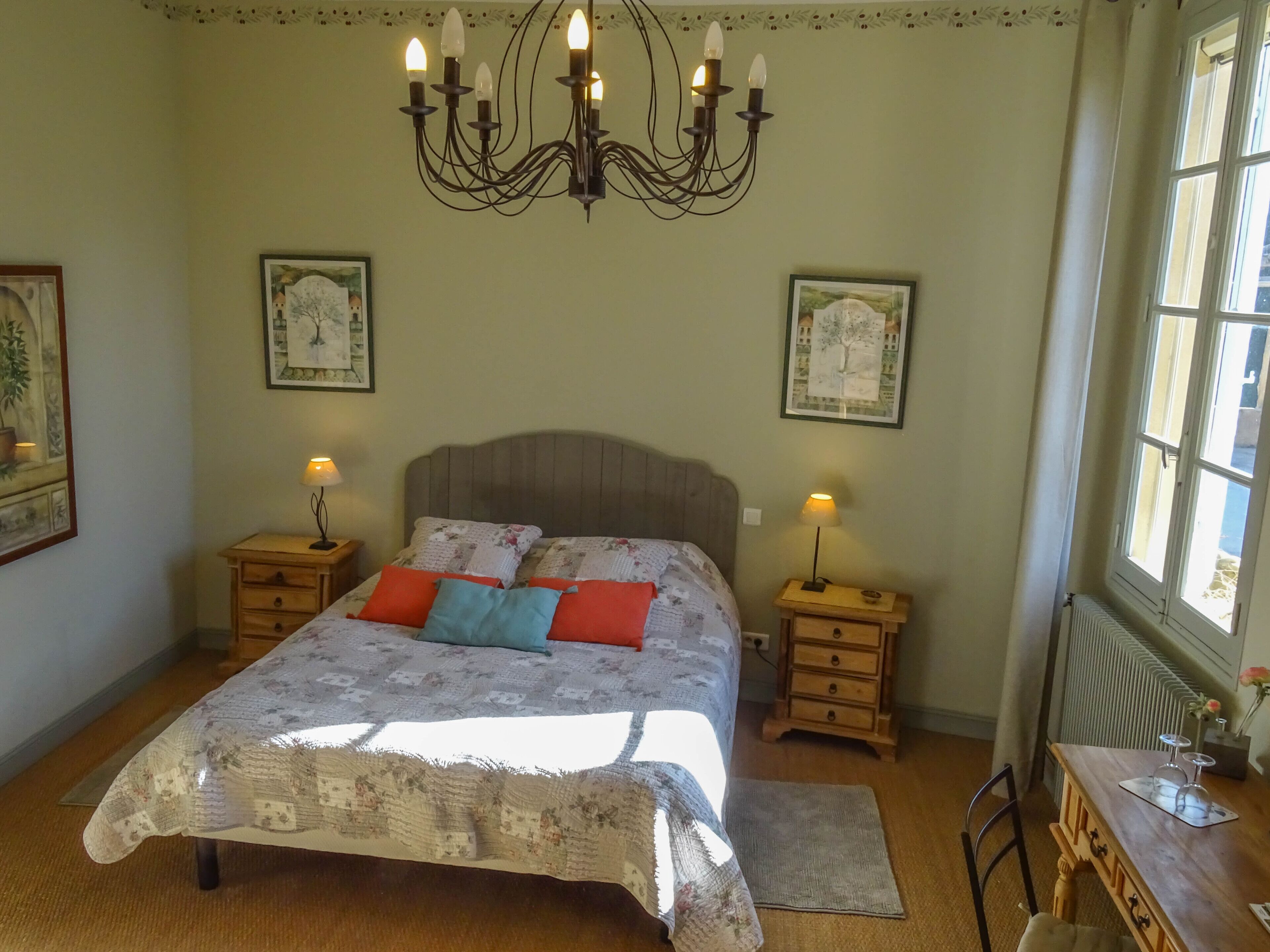 A bedroom with a large bed, two bedside tables with lamps, a chandelier, and framed pictures on the walls.
