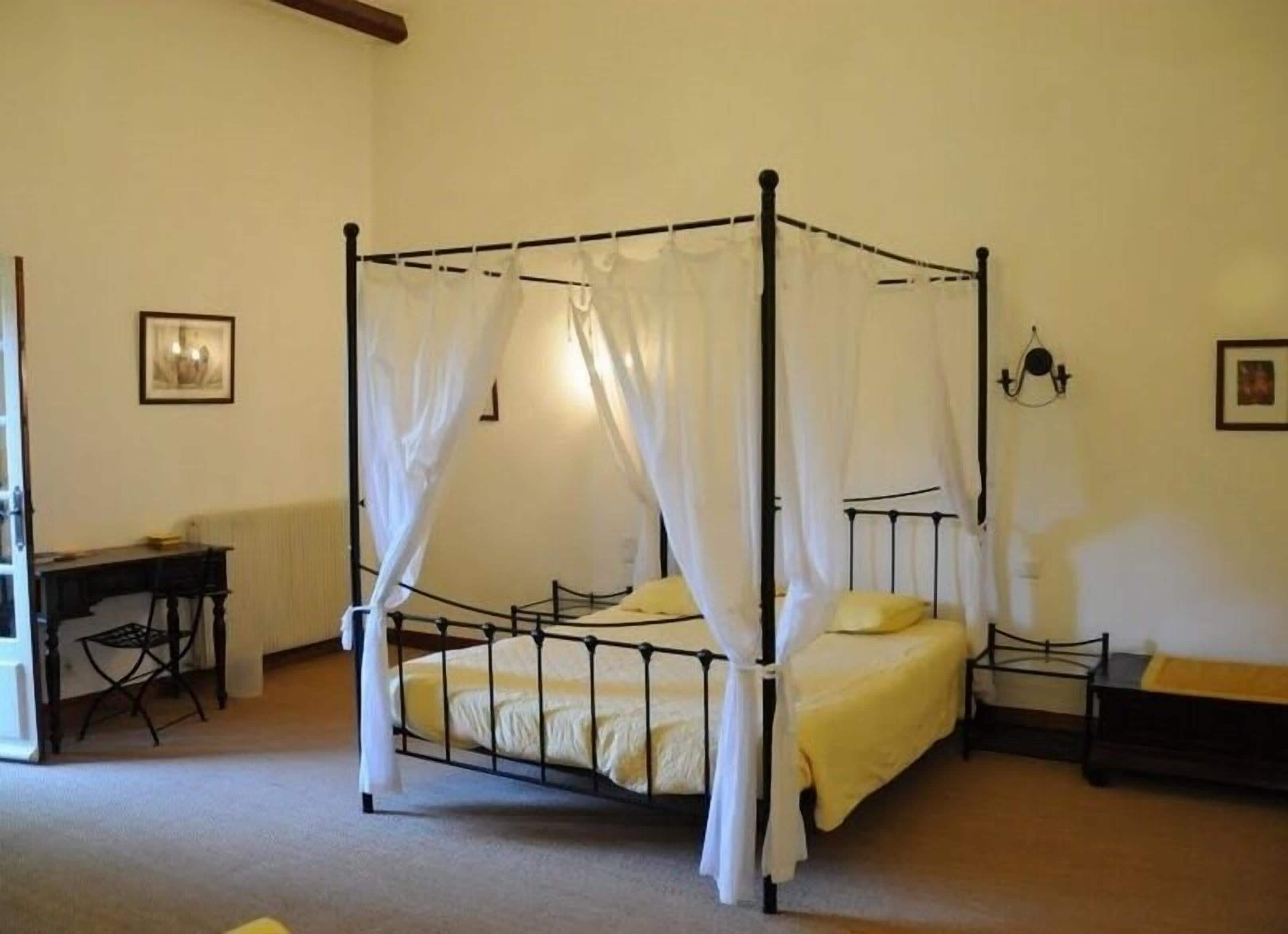 A four-poster bed with white curtains, a bedside table, and a chair.