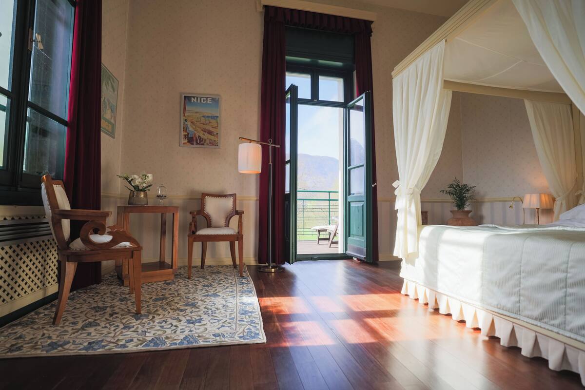 A bedroom with a canopy bed, a wooden floor, a small table with chairs, and a view through an open door.