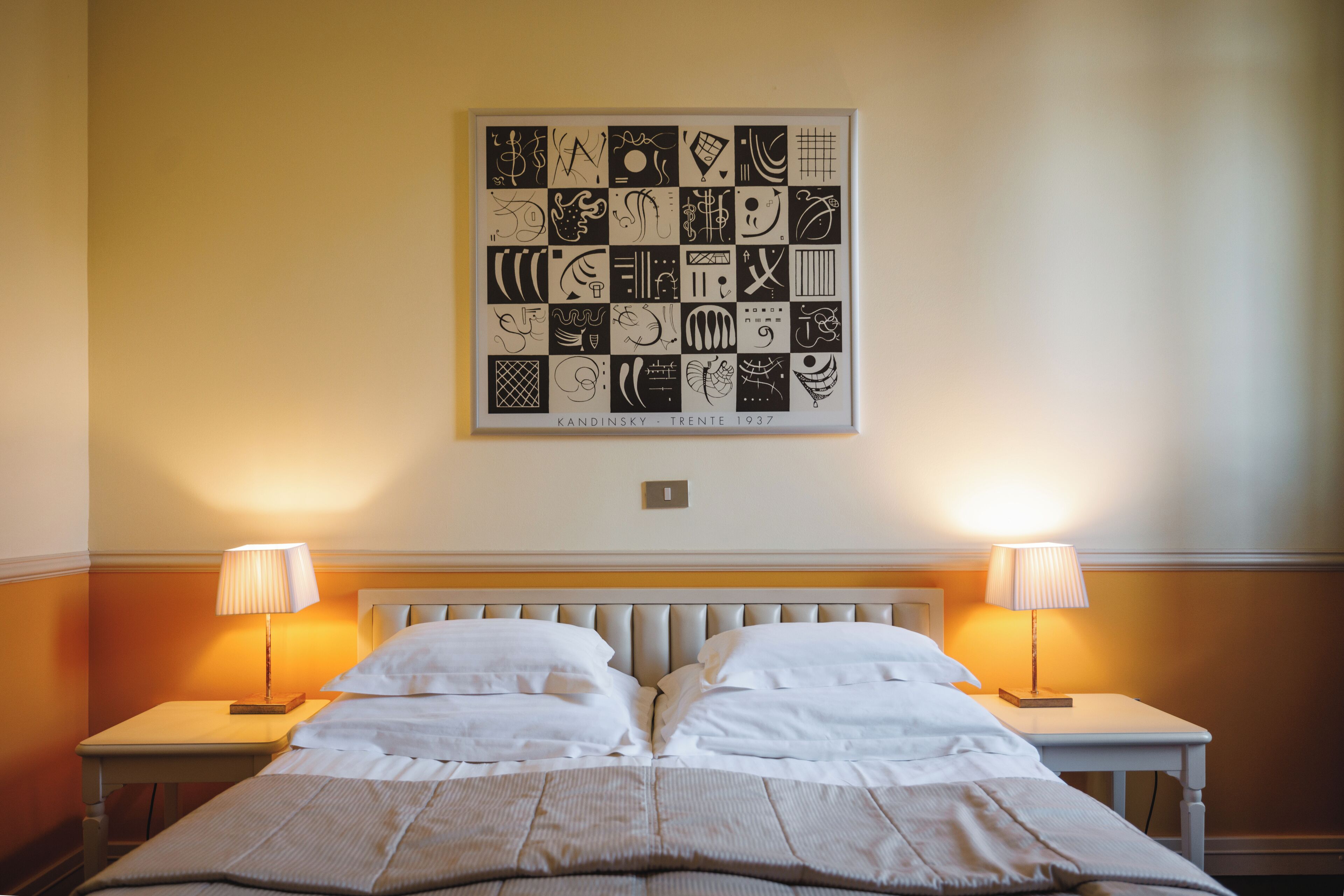 A hotel room with a neatly made bed, two bedside lamps, and a framed abstract art piece on the wall.