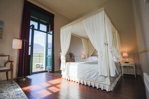 Una cama con dosel sobre cuatro postes con ropa de cama blanca, una silla de madera, una mesita con una lámpara y vistas al exterior a través de un ventanal.